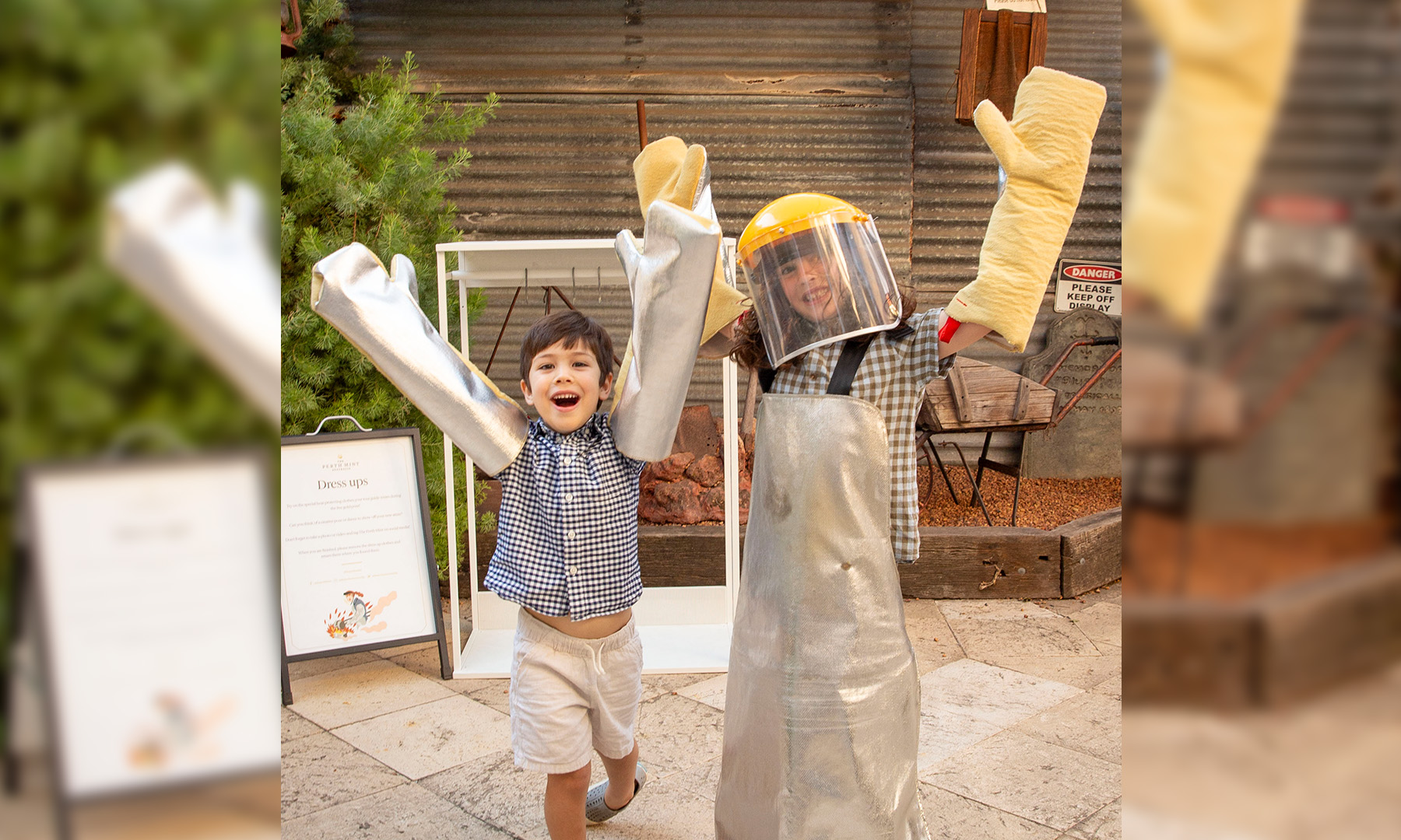 Two children in an outdoor courtyard dressed up in personal protective equipment, long gloves, apron, face visor