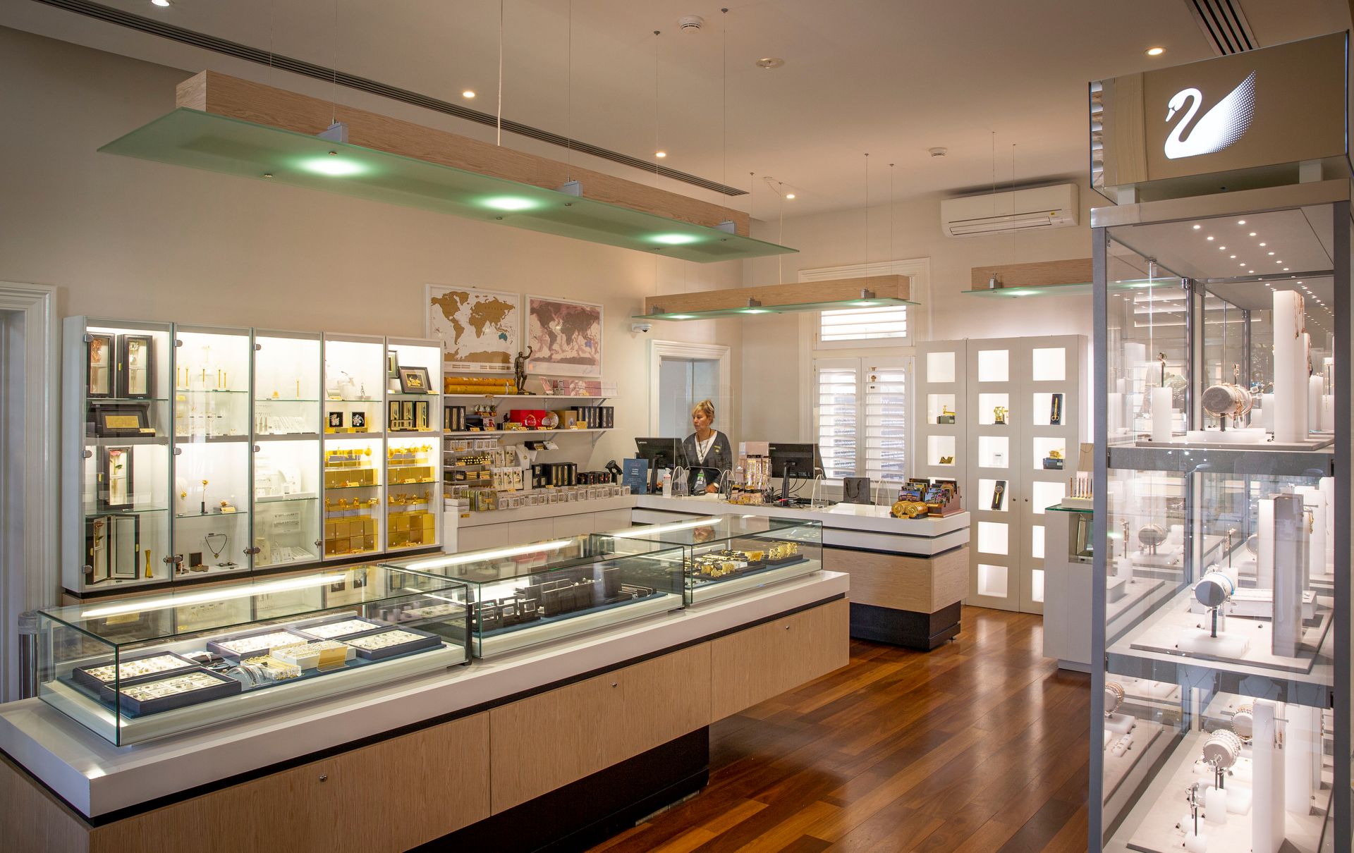 A shop with display cases and shelving along the walls and lower island display cases in the centre of the room containing jewellery and other souvenir items, a staff member stands behind a counter at the back of the room.