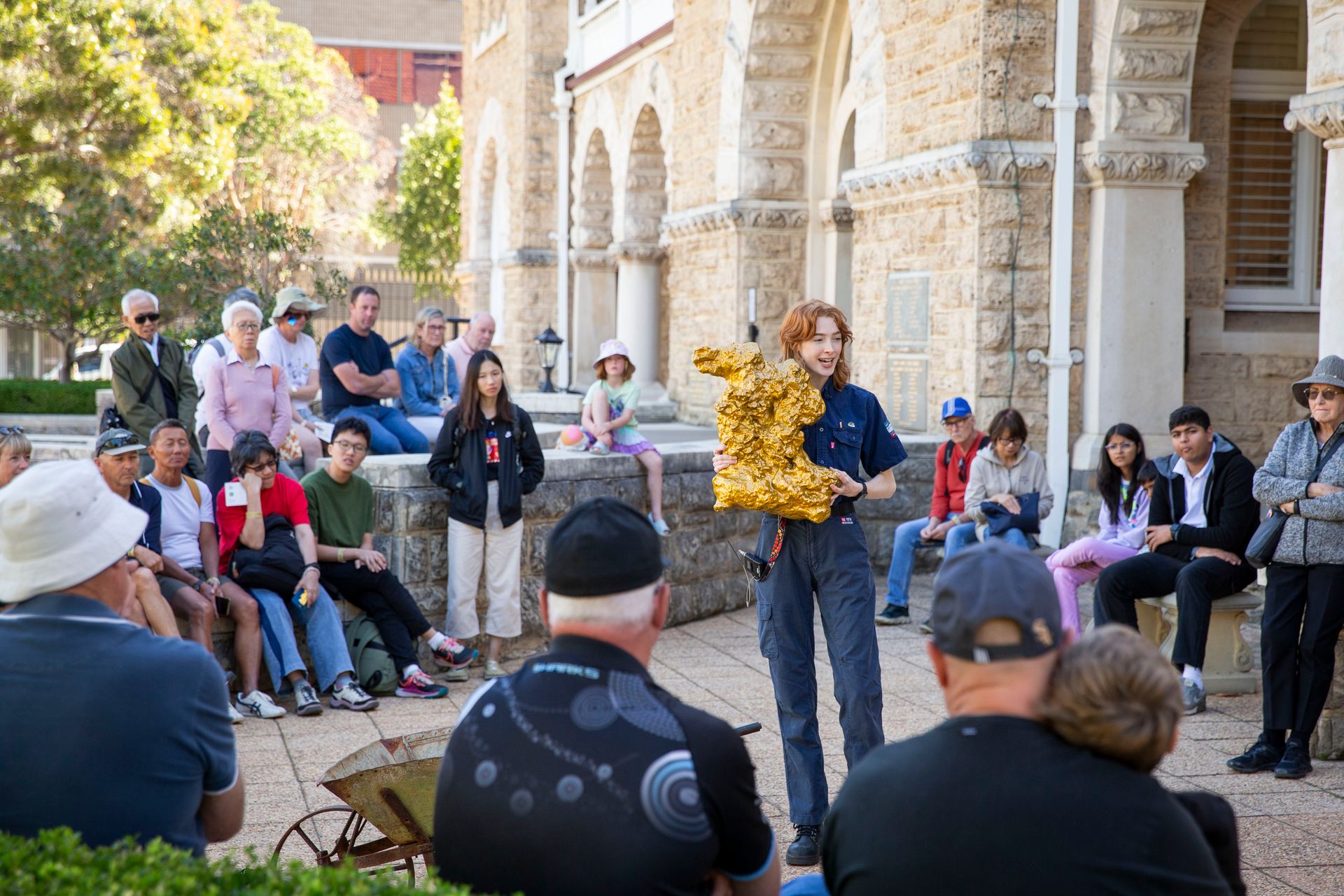 A tour guide holds a large replica gold nugget and is talking to a groups of visitors in the forecourt in front of a large limestone building