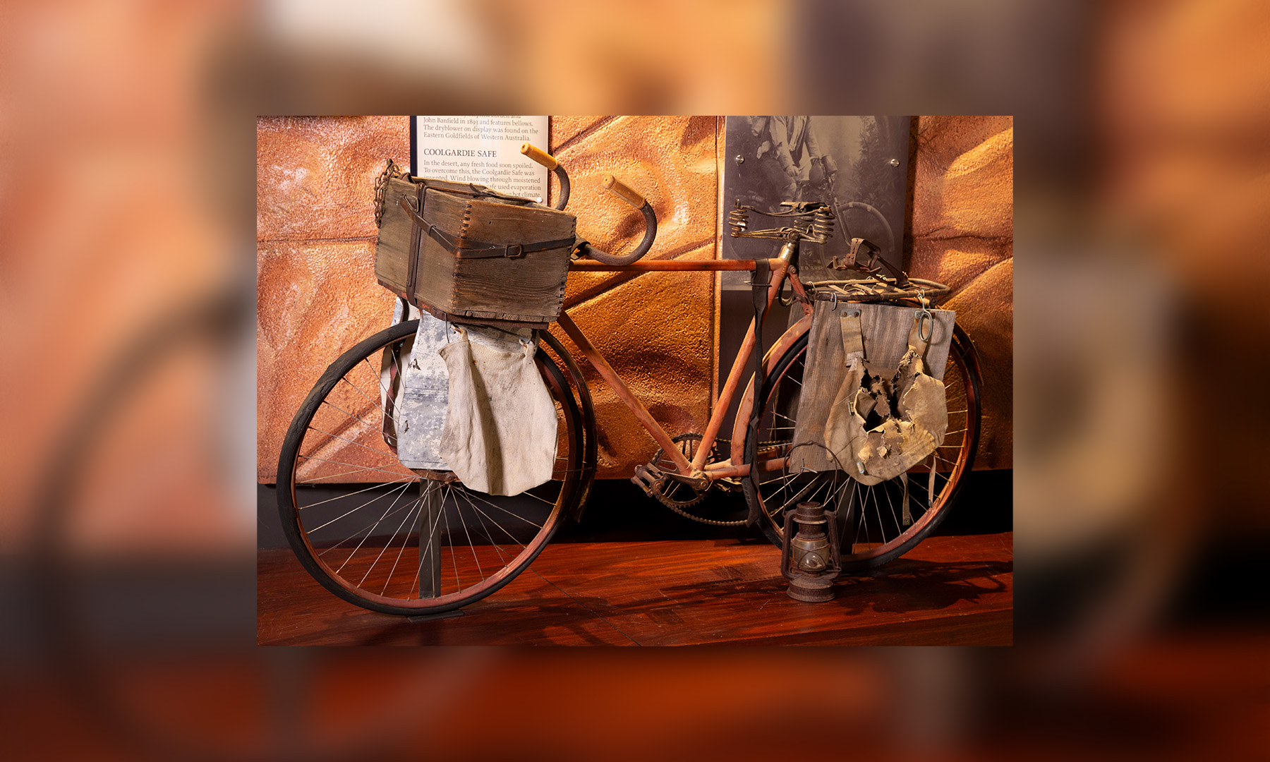An old bicycle with wooden crate fixed in front of handlebars and various hessian bags, a lantern and a rabbit trap hanging from the frame is on display in a museum gallery