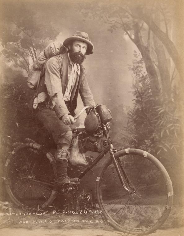 Sepia studio photograph of a man with a swag on his back sitting astride a bicycle with well worn tyres and that has mug, water bottle, hessian water bag and other items hanging from the handlebars and bike frame.