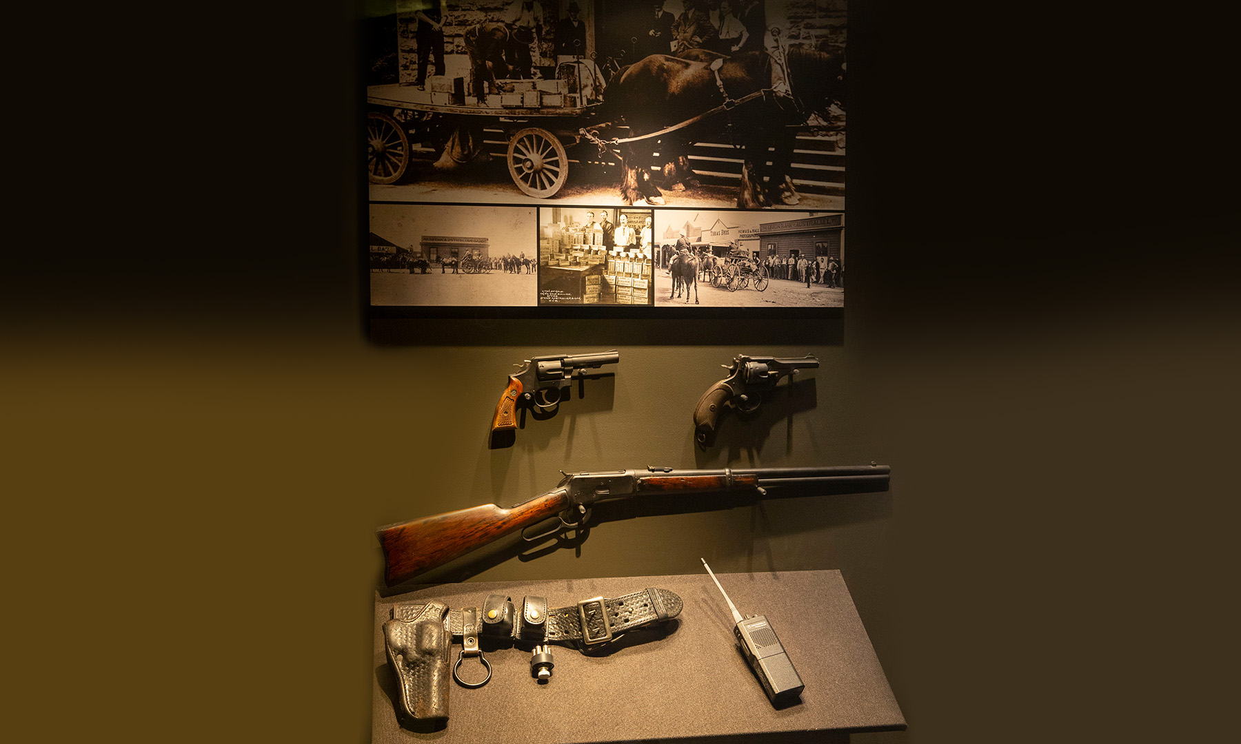 Museum display with historic black and white images of gold being delivered by horse and cart from the goldfields to The Perth Mint. Below are two revolvers, a rifle, a security gun belt and a walkie-talkie