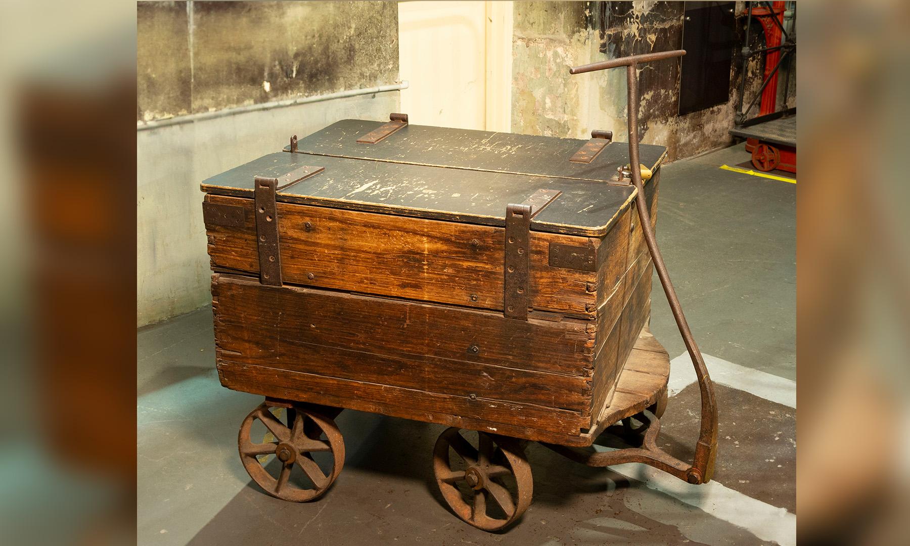 A late nineteenth century wooden cart with two double-hinged wooden lid, metal wheels and long metal handle sits in industrial factory