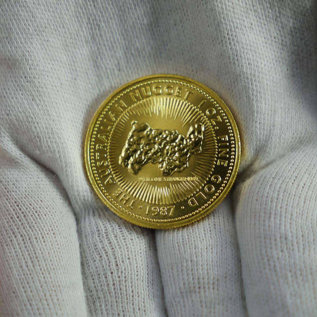 A gold coin 'The Australian Nugget 1 oz. Fine Gold 1987' with a design of the 'Welcome Stranger' gold nugget encircled by rays of varying lengths is held in the palm of a hand wearing a white glove
