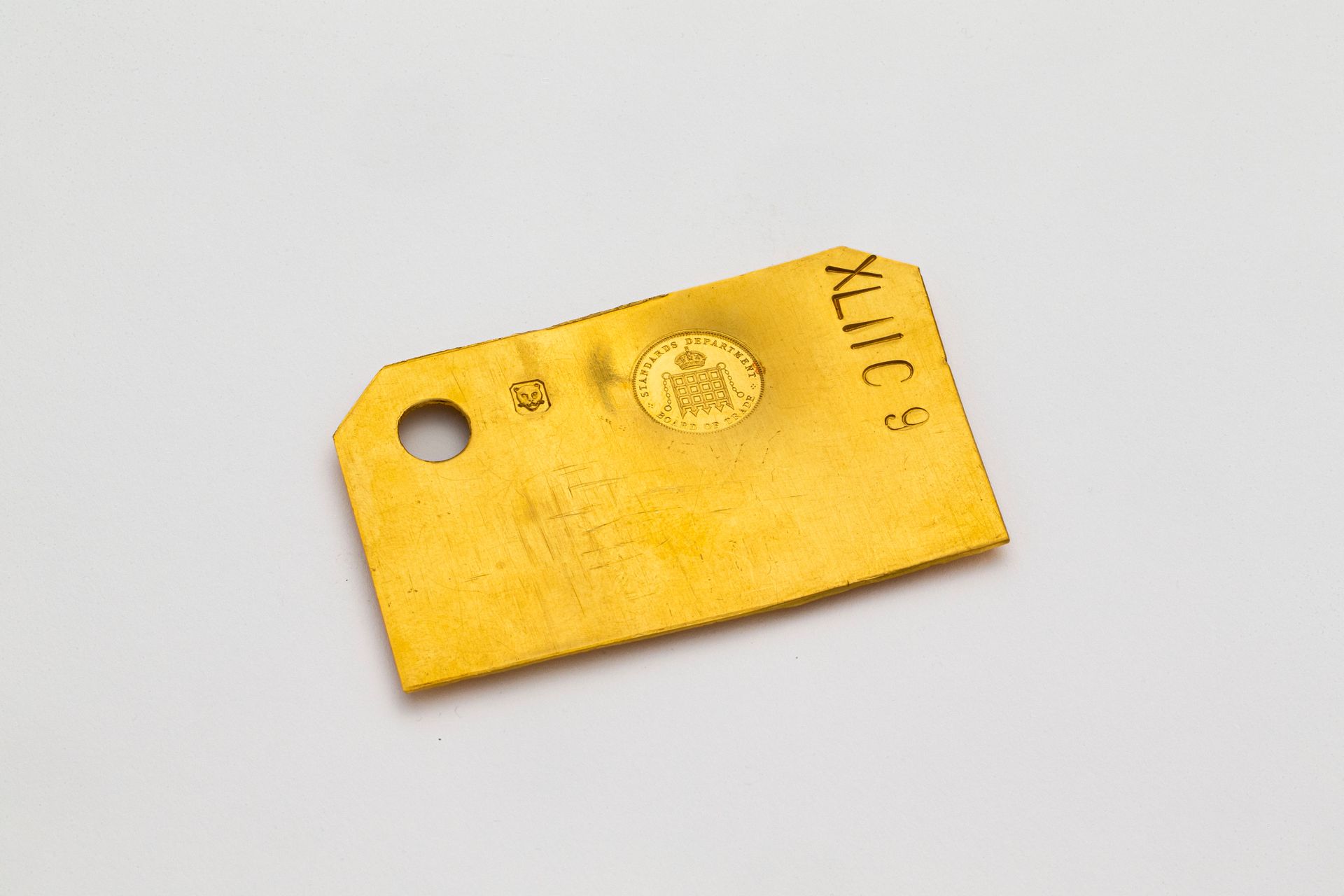 A rectangular gold plat with the top left corner cut off. Roman numerals 'XLIIC 9' and a stamp with a crown over a portcullis 'Standards Department Board of Trade', also the stamp of the London Assay Office