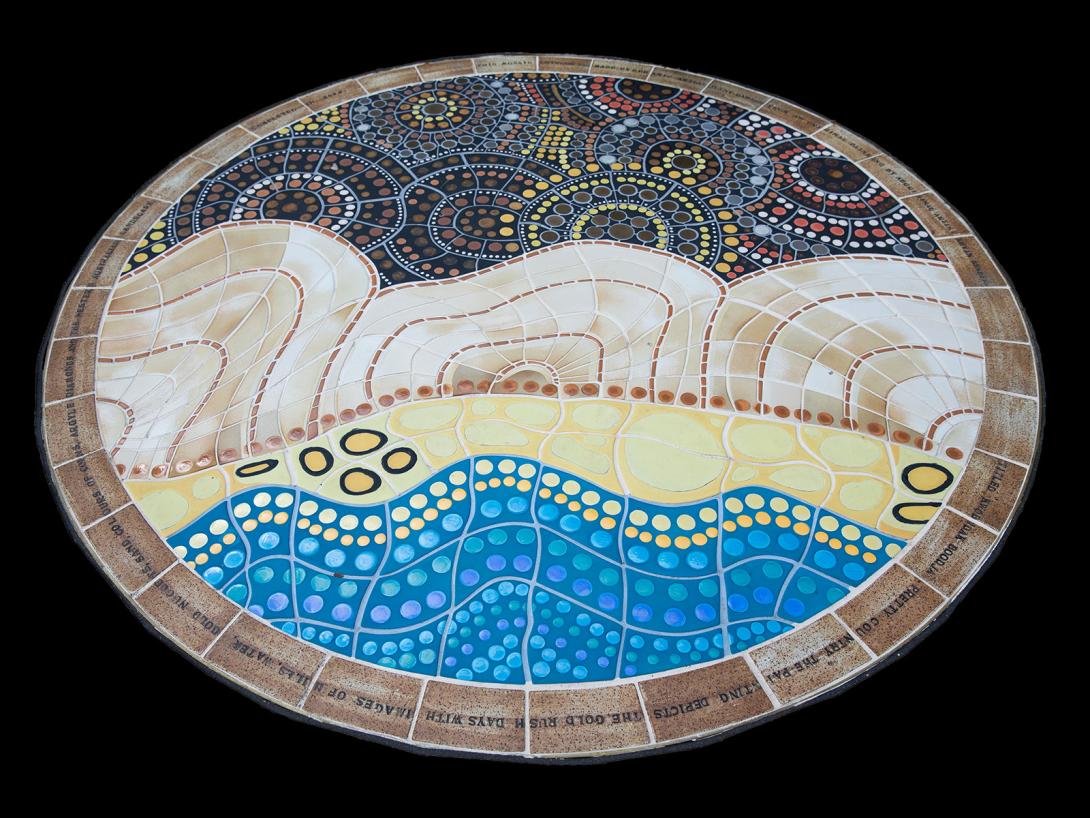 A circular mosaic artwork inlaid on the ground made of small tiles - the lower section has a design of blue and yellow tiles that depicts the gold rush days with images of hills, water, sand, and golden nuggets. The upper part of the mosaic includes shades of gold and silver - depicting coins.