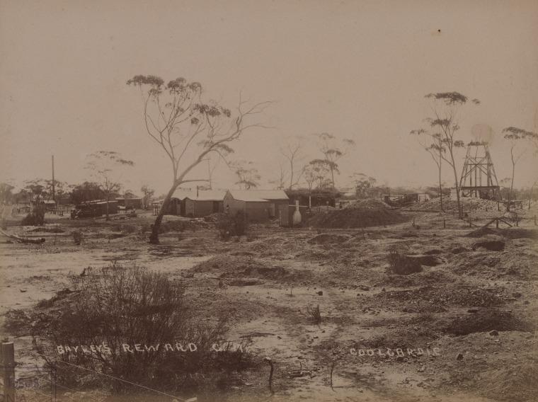 Sepia image circa 1895 titled 'Bayley's Reward Gold Mine, Coolgardie' showing arid landscape with numerous dirt mounds, several trees, corrugated iron buildings in the centre of the image and to the right a metal mine headframe.