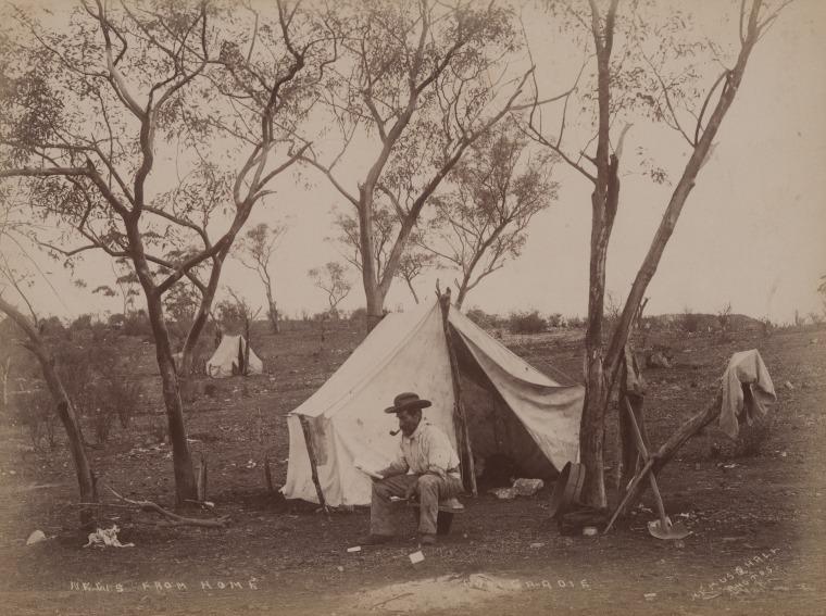 Sepia photograph circa 1895 titled 'News from home, Coolgardie', image of a man sitting on a makeshift chair of a bucket and gold pan in front of a white canvas tent with another tent in the background; bushland with sparse trees; man is smoking a pipe and reading.