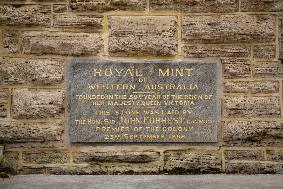 A stone plaque inset into a limestone wall reads 'Royal Mint of Western Australia founded in the 59th year of the reign of Her Majesty Queen Victoria. This stone was laid by the Hon. Sir John Forrest, K.C.M.C. Premier of the Colony 23rd September 1896'