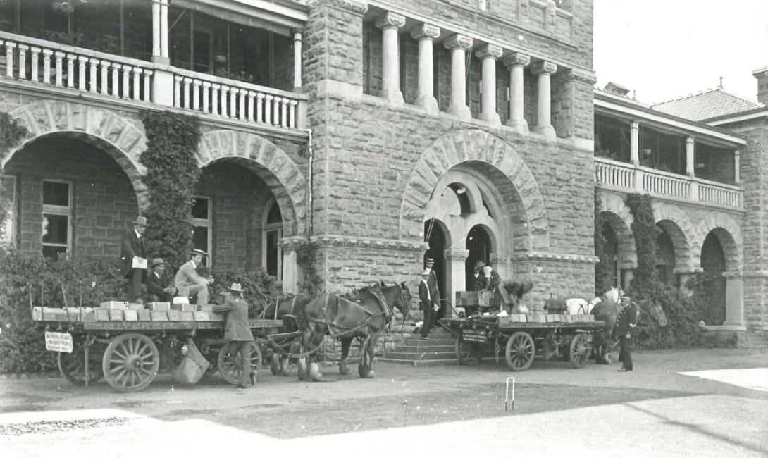 Two wagons, each drawn by two horses, are stopped in front of a two story building with a limestone facade. The wagons are laden with wooden boxes which are being unloaded by two labourers onto an open top wooden wagon at the front double arched doors of the building. A police officer stands guard.