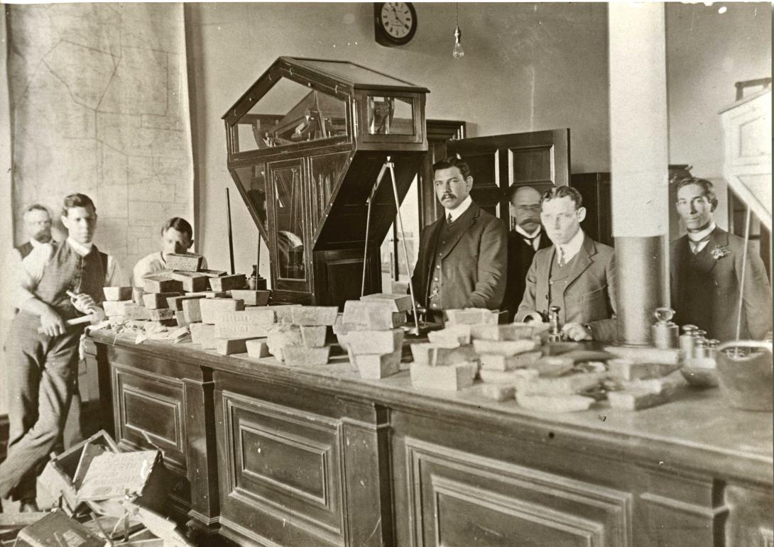 Black and white image of a group of workers dressed in suits standing inside an office around a long wooden counter with a very large set of scales to weigh gold bullion, which is laid out across the length of the counter
