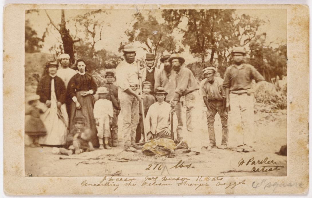 Black and white photograph 'Unearthing the Welcome Stranger Nugget', 1869, photographer W. Parker; image shows a group of miners, their wives and children posing with the finders of the nugget, Richard Oates, John Deason - both holding picks - and his wife. The stone in the foreground is not the gold nugget but a rock