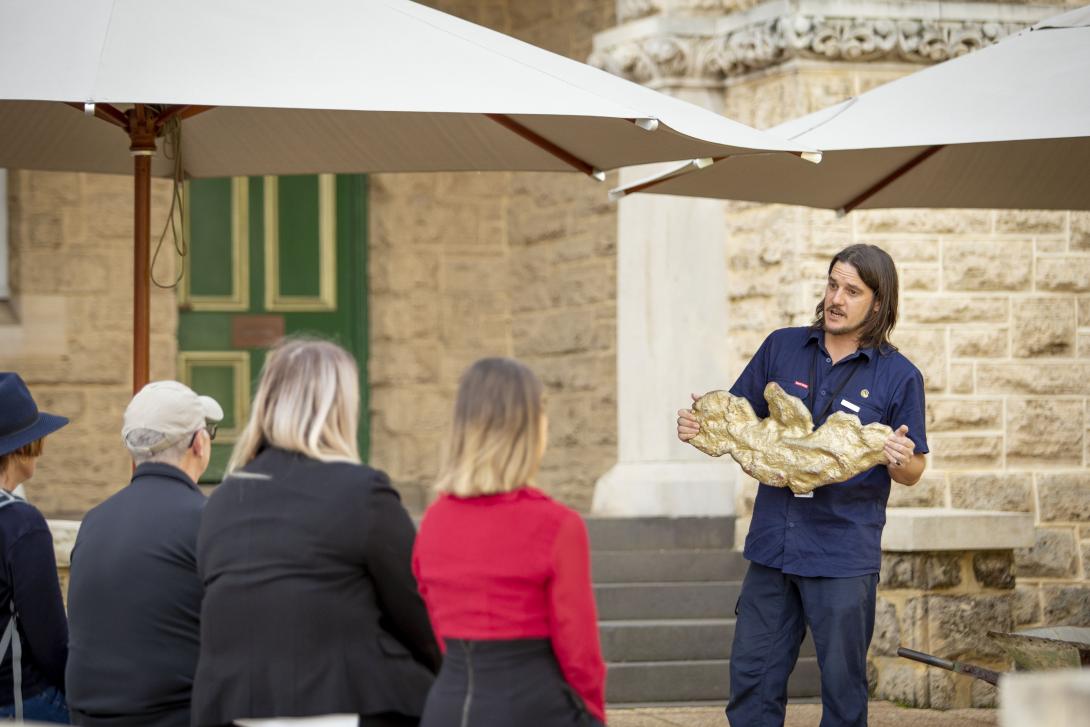 Visitors is outside a building with a limestone facade listening to a tour guide in a blue uniform who is holding a replica of the 'Golden Eagle' gold nugget, which is shaped like a bird with wings outstretched.