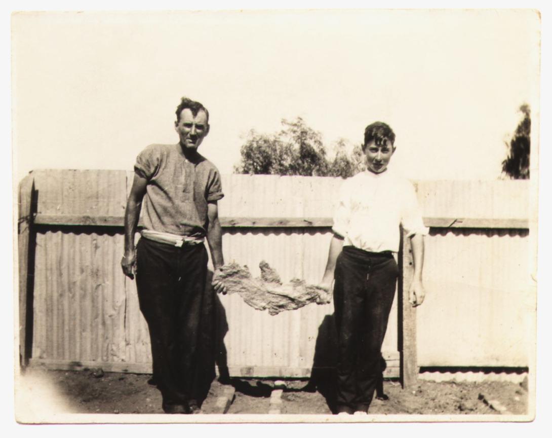 Black and white image of 16 year old Jimmy Larcombe and his father, Jim senior standing in front of a corrugated iron fence, each holding one end of the large gold 'Golden Eagle' nugget.