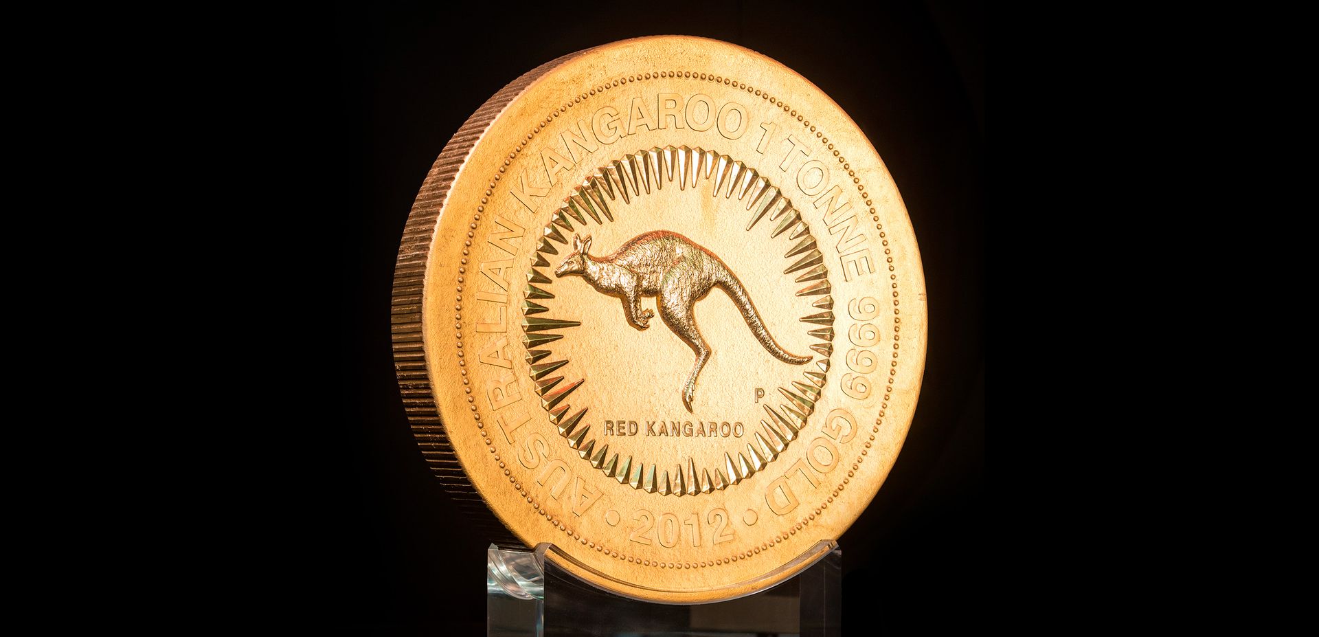 A large gold coin 'Australian Kangaroo 1 Tonne 9999 Gold 2012' with central design of jumping red kangaroo inside a circle of rays is supported on an acrylic mount.