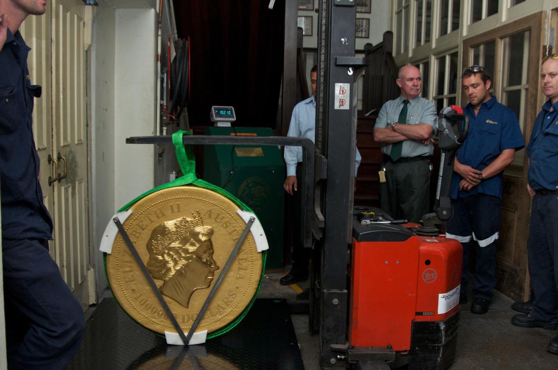 A huge gold coin with the portrait of Queen Elizabeth visible is suspended from a forklift and has been lowered onto a large scale which has a reading of 1021.0kg, five men stand around obvserving.
