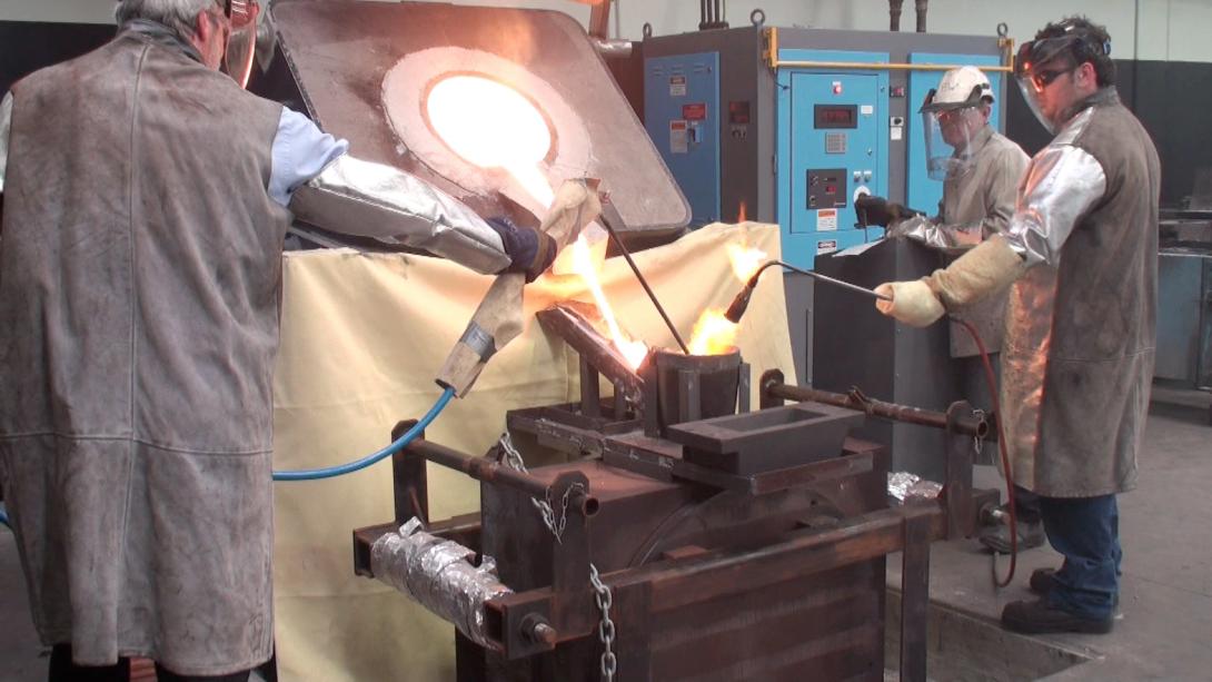 A large cast iron mould is positioned below a huge industrial furnace and red hot molten gold is being poured from the furnace into the opening of the mould, three workers wearing safety aprons, gloves and face visors are operating various pieces of machinery, one hold a blowtorch above the opening of the mould.