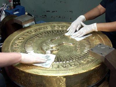 A large gold coin lying with the reverse surface visible, which has the design of a jumping red kangaroo surrounded by a circle of rays, is being polished by two people wearing white golves using white cloths