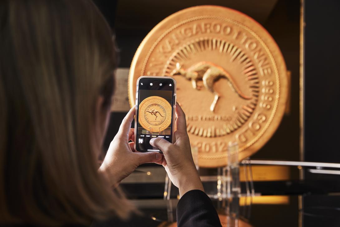 A woman holding a mobile phone is taking a photograph of a huge gold coin 'Australian Kangaroo 1 Tonne 9999 Gold 2012' that is supported by an acrylic plinth in an exhibition gallery.