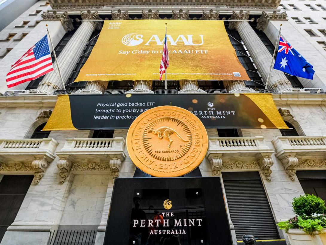 A huge gold coin with a design of a jumping kangaroo encircled by rays 'Australian Kangaroo 1 Tonne 999 Gold 2012' is on display on a black plinth labelled 'The Perth Mint Australia' outside the New York Stock Exchange, a tall building with ornate columns and balconies built into the facade.