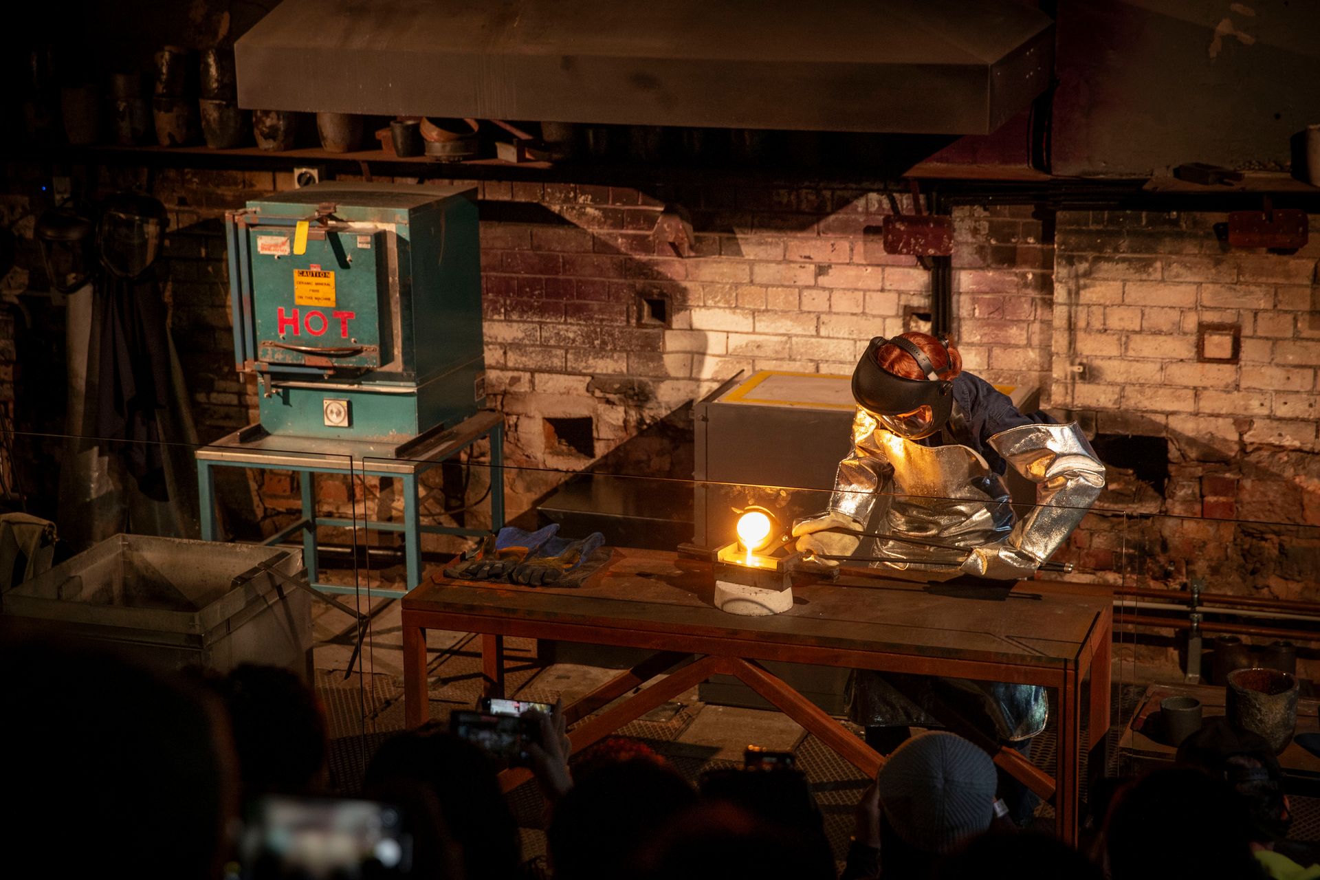 Worker wearing protective safety equipment is holding a red hot crucible with long tongs and tipping the crucible to pour molten gold into a cast iron rectangular mould