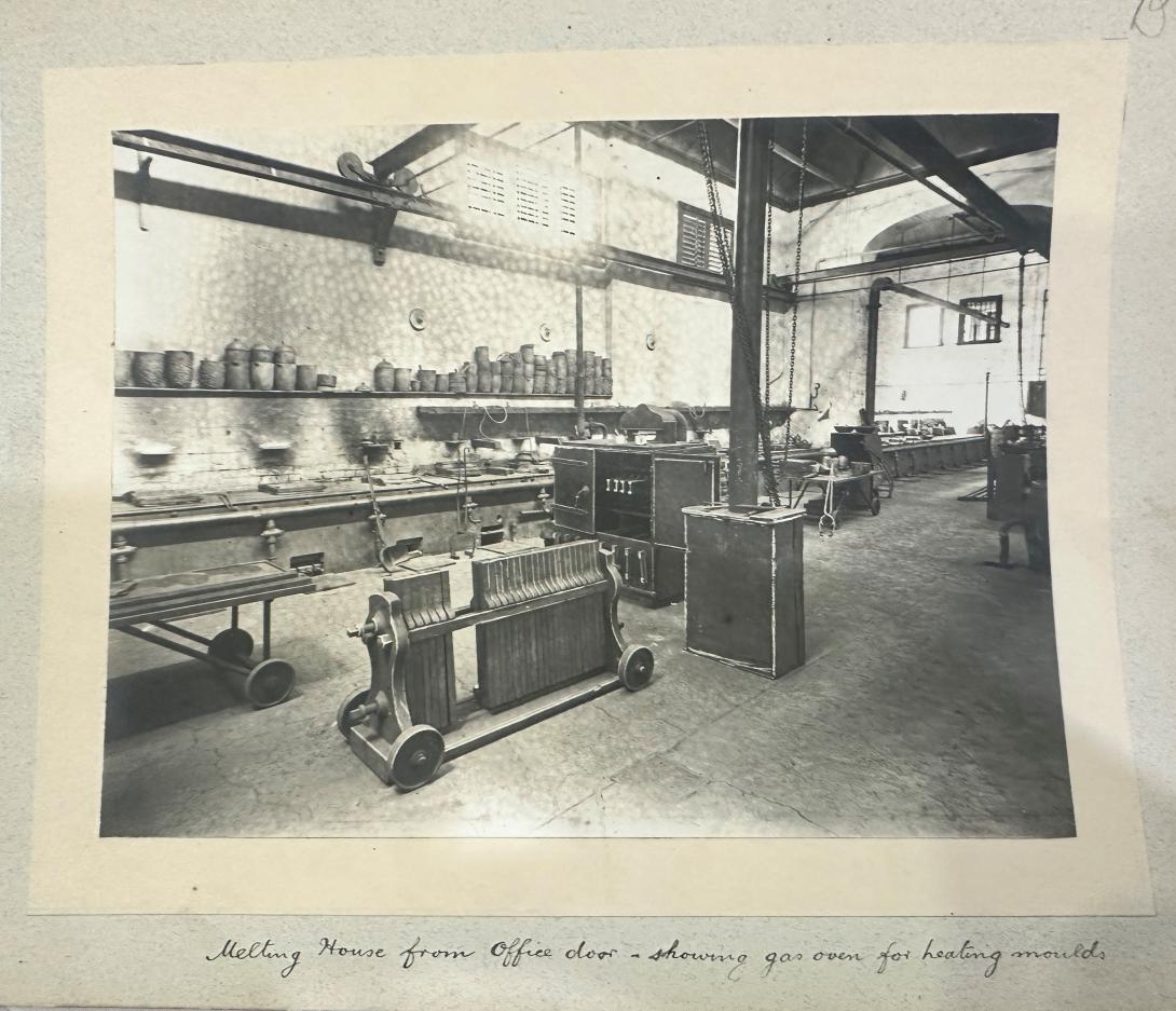 Black and white photo stuck into and 1908 album titled 'Melting House from Office door - showing gas oven for heating moulds' - photo is a factory room with back wall lined with a row of furnaces above which is a shelf stacked with clay and graphite crucibles, in the foreground is a trolley from which cast iron moulds are hanging, in the centre of the room is a low table with wheels and an oven containing some moulds.