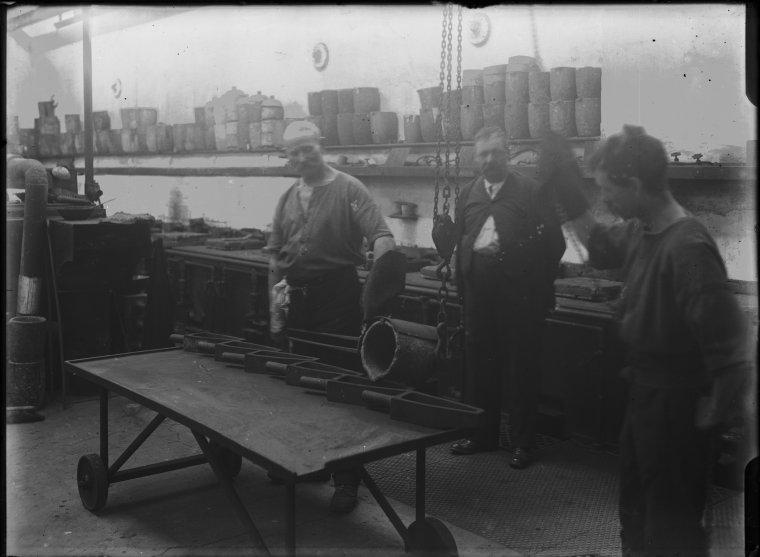 Black and white photo of three workers standing inside a factory room with furnaces along the back wall and a shelf full of crucibles above and a low table on wheels in the foreground.