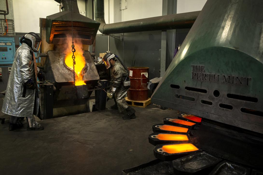 Interior of industrial factory with large furnace and fume extraction hood - molten gold is being poured from the furnace into a crucible by two men dressed in full personal protective equipment including helmets and kevlar jackets, gloves and spats; a machine with a circle of gold bar moulds is in the foreground, several have been filled with gold