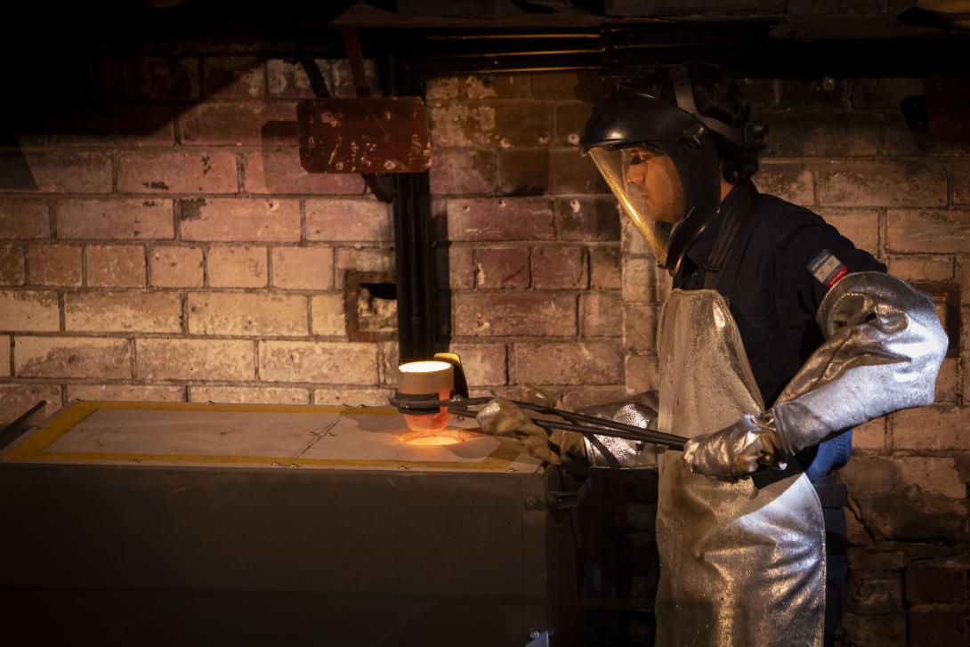 A worker dressed in personal protective gear - face shield, long silver kevlar gloves and apron, lifts a red hot crucible containing molten gold out of a furnace with a pair of iron lifting tongs