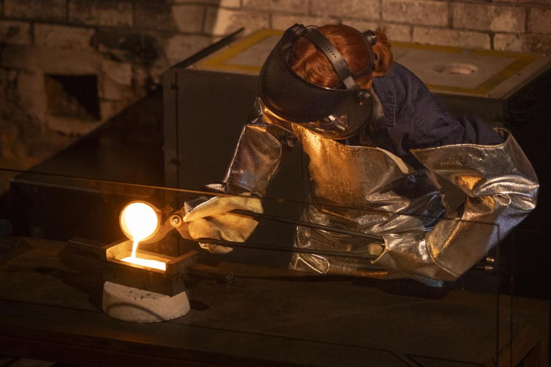 Worker wearing protective safety equipment is holding a red hot crucible with pouring tongs and tipping the crucible to pour molten gold into a cast iron rectangular mould