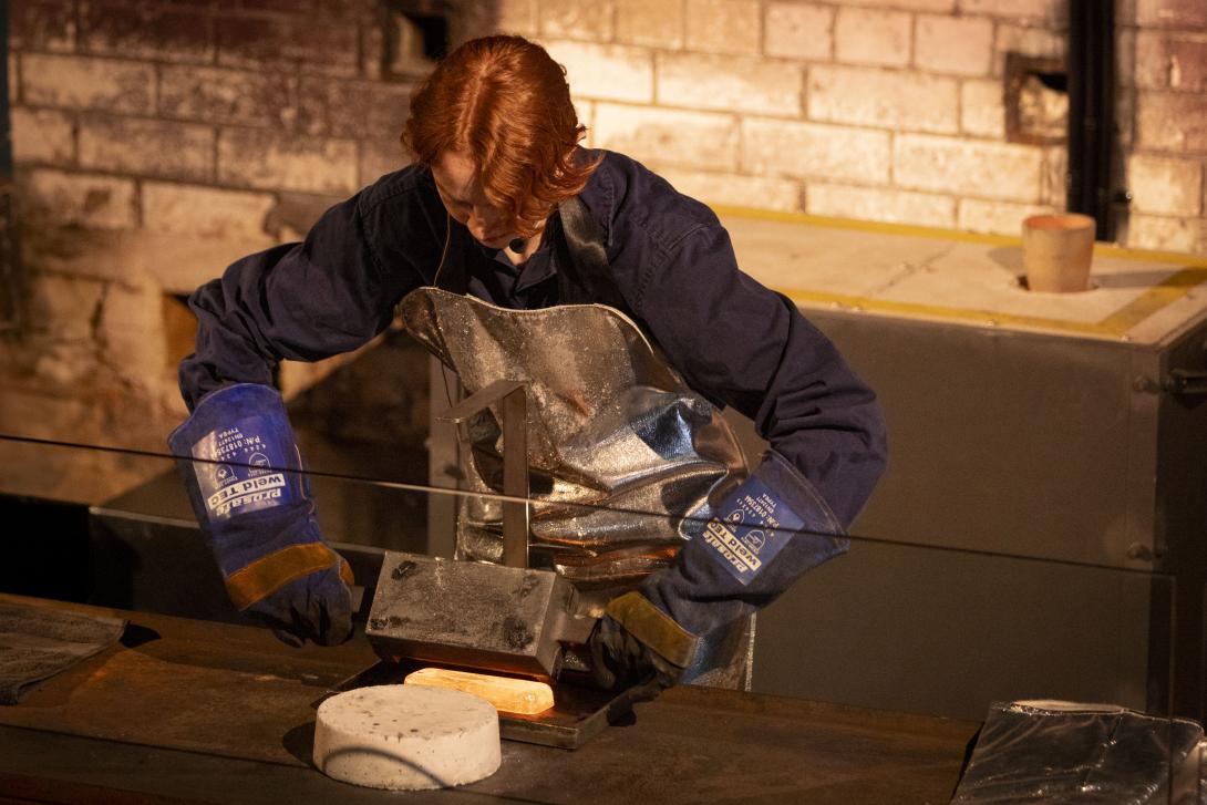 Worker wearing blue uniform and silver kevlar tips a hot cast gold bar out of a mould onto a tray