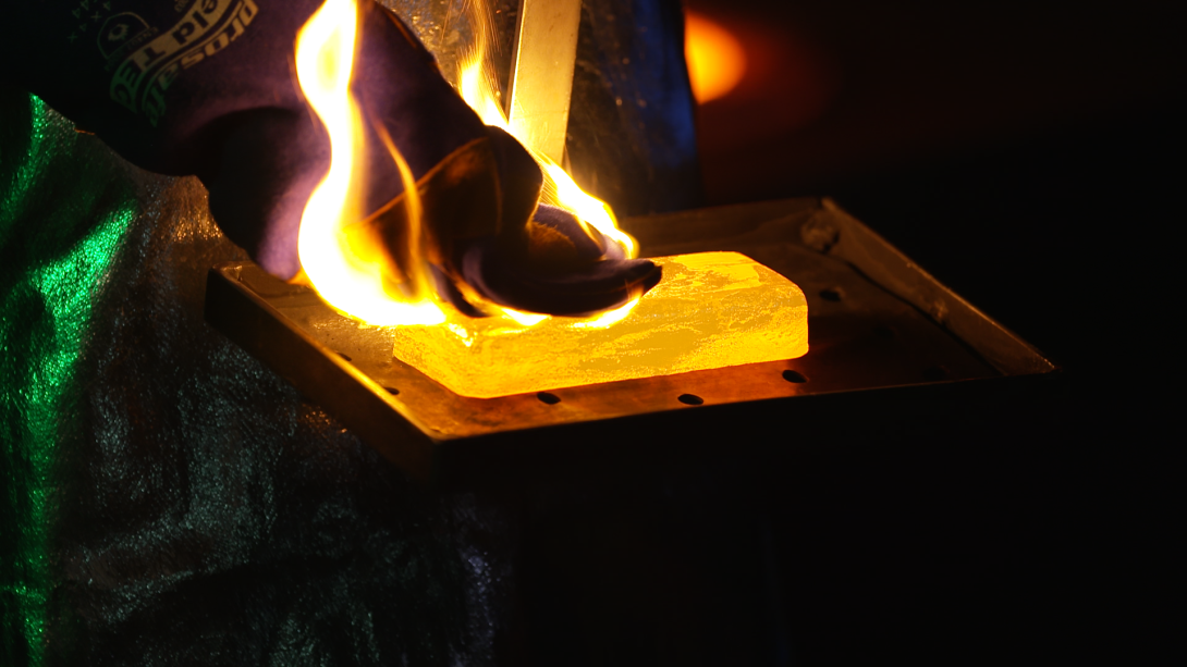A gloved hand is resting on a red hot cast gold bar that is positioned on a tray; the gold bar has flames coming out the top and around the glove