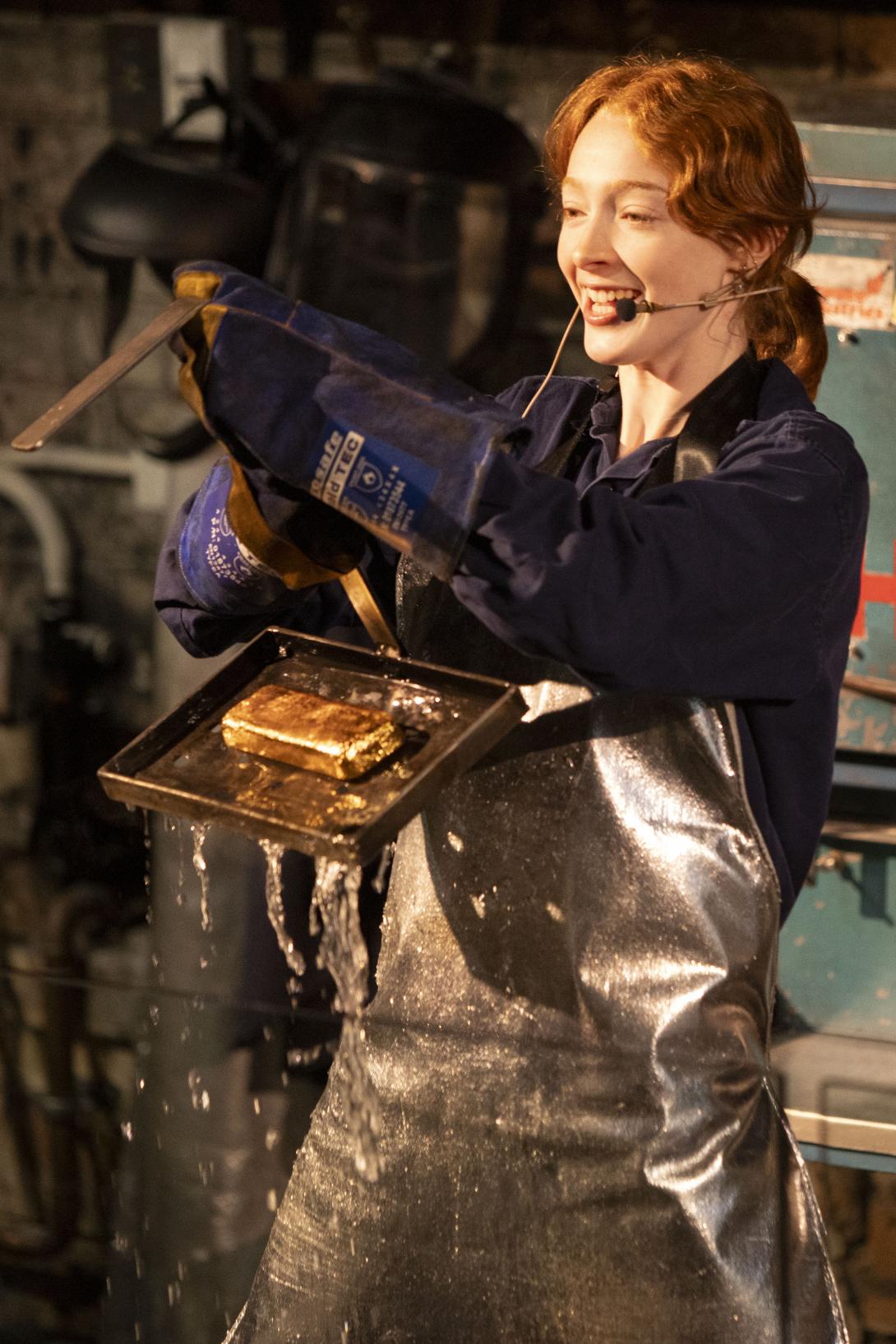 Worker wearing blue uniform and silver kevlar pulls a cast gold bar on a tray up out of a tank of water a mould onto a tray