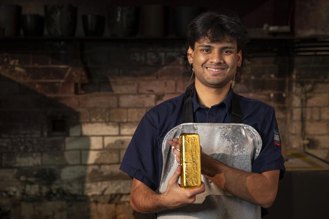 A worker in a blue uniform and silver kevlar apron holds up a cast gold bar, the work is standing inside a melting house, a furnace is in the background.