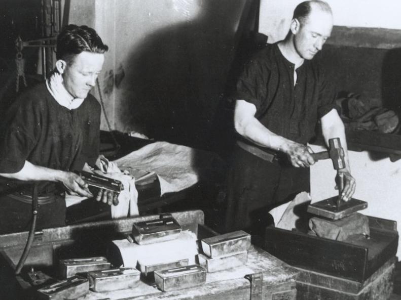 Two workers in a factory stand near a wooden cart laden with cast gold bars; one worker is holding a gold bar while the second worker holds a metal stamp and a mallet which he is using to stamp a gold bar which has a jig placed on top of it to ensure accurate placement of the stamp