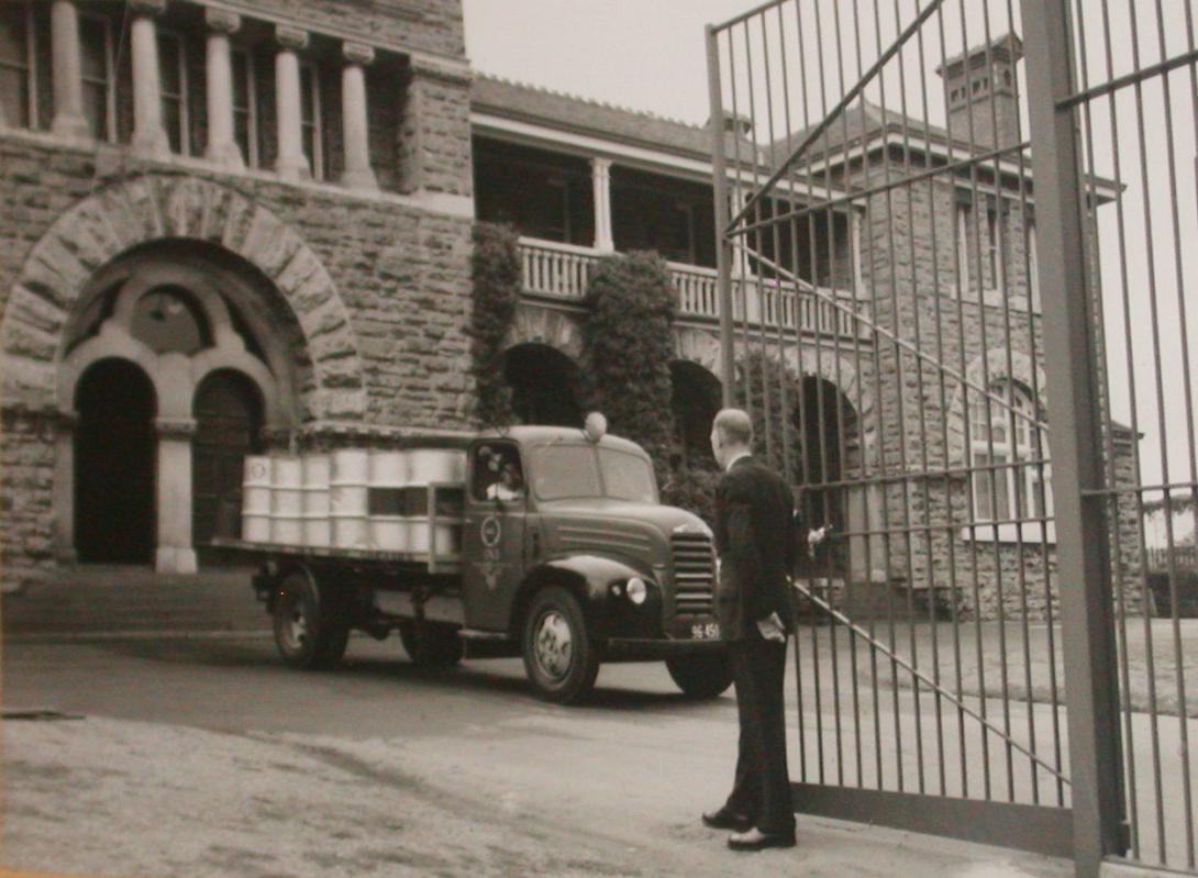 An open tray truck with a load of 44 gallon drums is positioned in from of a limestone clad building with and arched entrance and two arched entrance doors. The back of a man in a suit is in the foreground. He stands holding open one half of a set of tall barred gates..