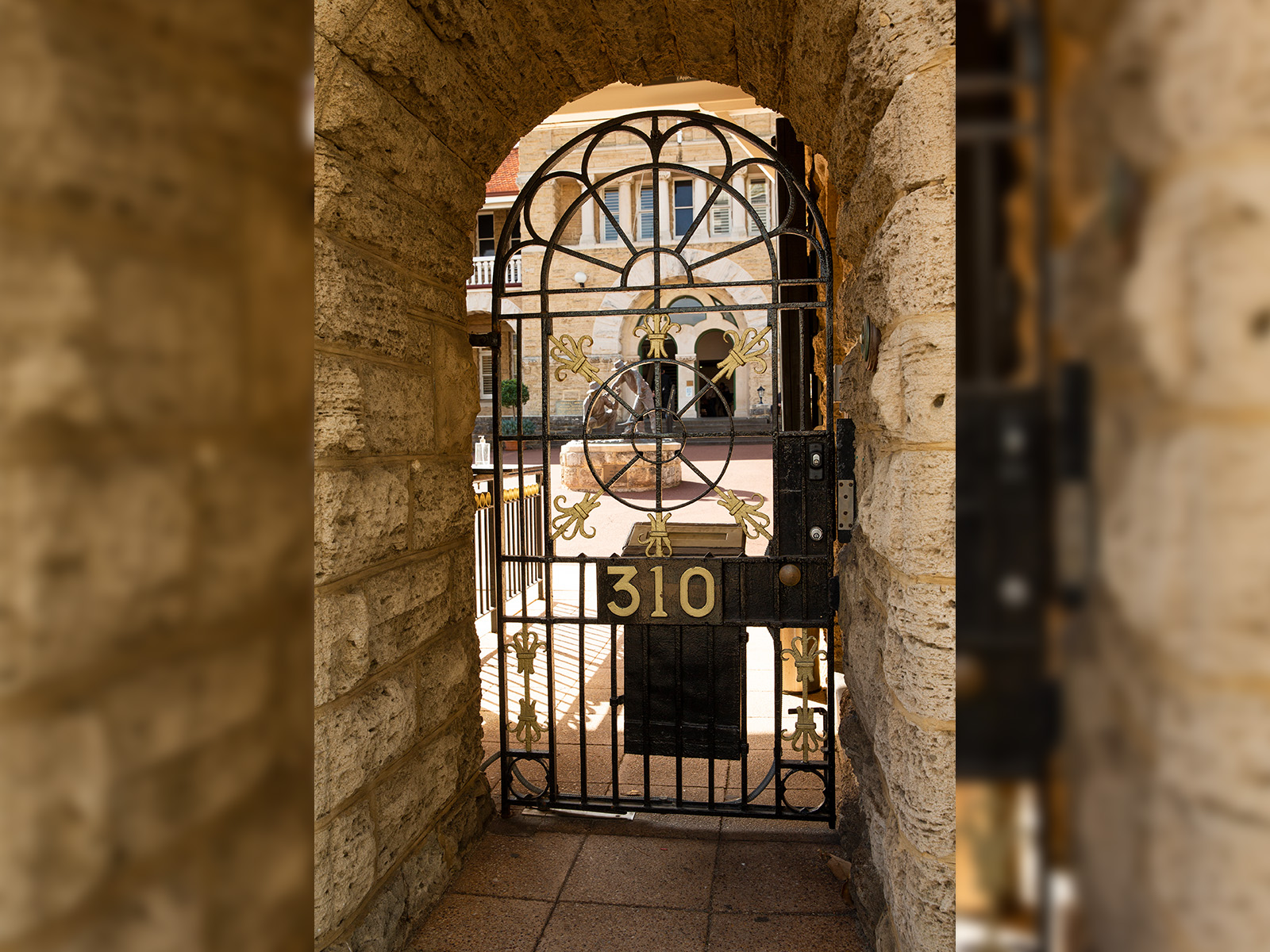 Limestone walls with and arched wrought iron pedestrian gate with gold number '310' and a black letter box; behind the gate is a building with limestone façade