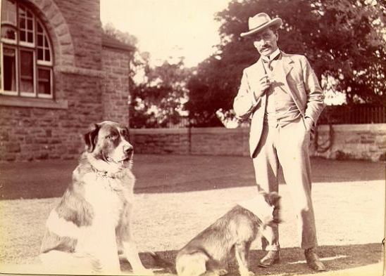 Sepia historical photo of man wearing three piece suit and hat with two large dogs, standing on lawn with limestone building behind