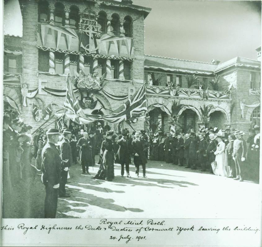 Black and white image of The Perth Mint, a two story building with three story central tower, limestone facade, decorated in bunting and Union Jack flag with the letters 'ER' and the British Coat of Arms positioned centrally above the double entrance doors. A large gathering of finely dressed people line either side of a path down which a group of people are walking
