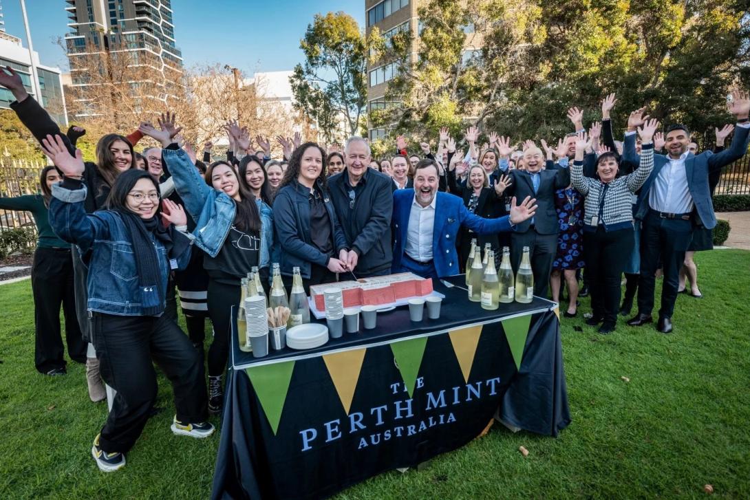 A group of employees, smiling and with hands in the air, stand around a table positioned outdoors on lawn; a birthday cake is being cut by two employees