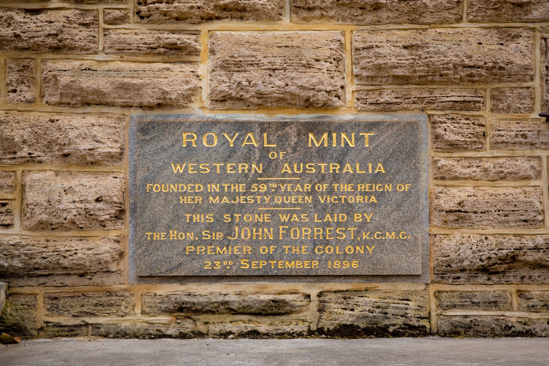 A stone plaque inset into a limestone wall reads 'Royal Mint of Western Australia founded in the 59th year of the reign of Her Majesty Queen Victoria. This stone was laid by the Hon. Sir John Forrest, K.C.M.C. Premier of the Colony 23rd September 1896'