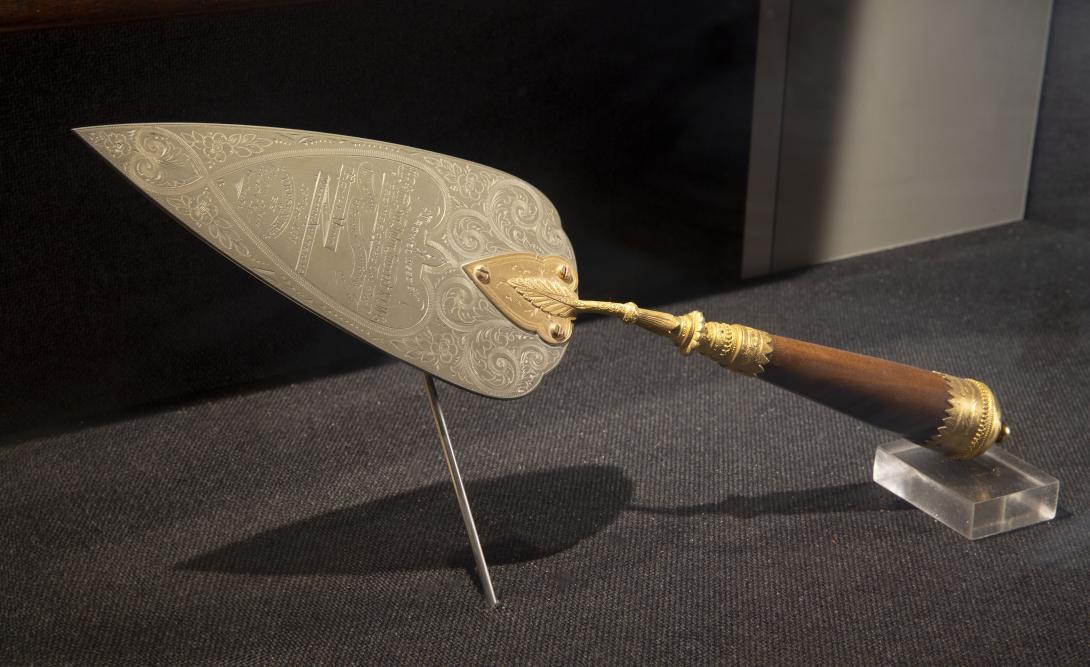 a metal presentation trowel with wood and gold handle is engraved 'Trowel used by the Hon Sir John Forrest KCMC "Premier" of the Colony in laying the Foundation Stone of the Royal Mint of Western Australia 23rd Sept. 1896. Presented by William Atkin, Robert O. Law Contractors.'