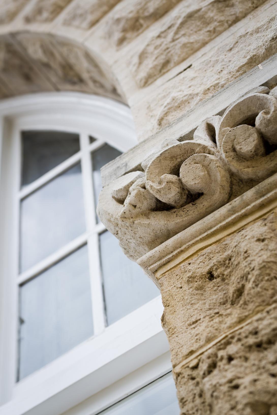 Detail of limestone clad building with portion of arched window with white timer frame and details to string course of limestone featuring carves swirl and stylised banskia nut