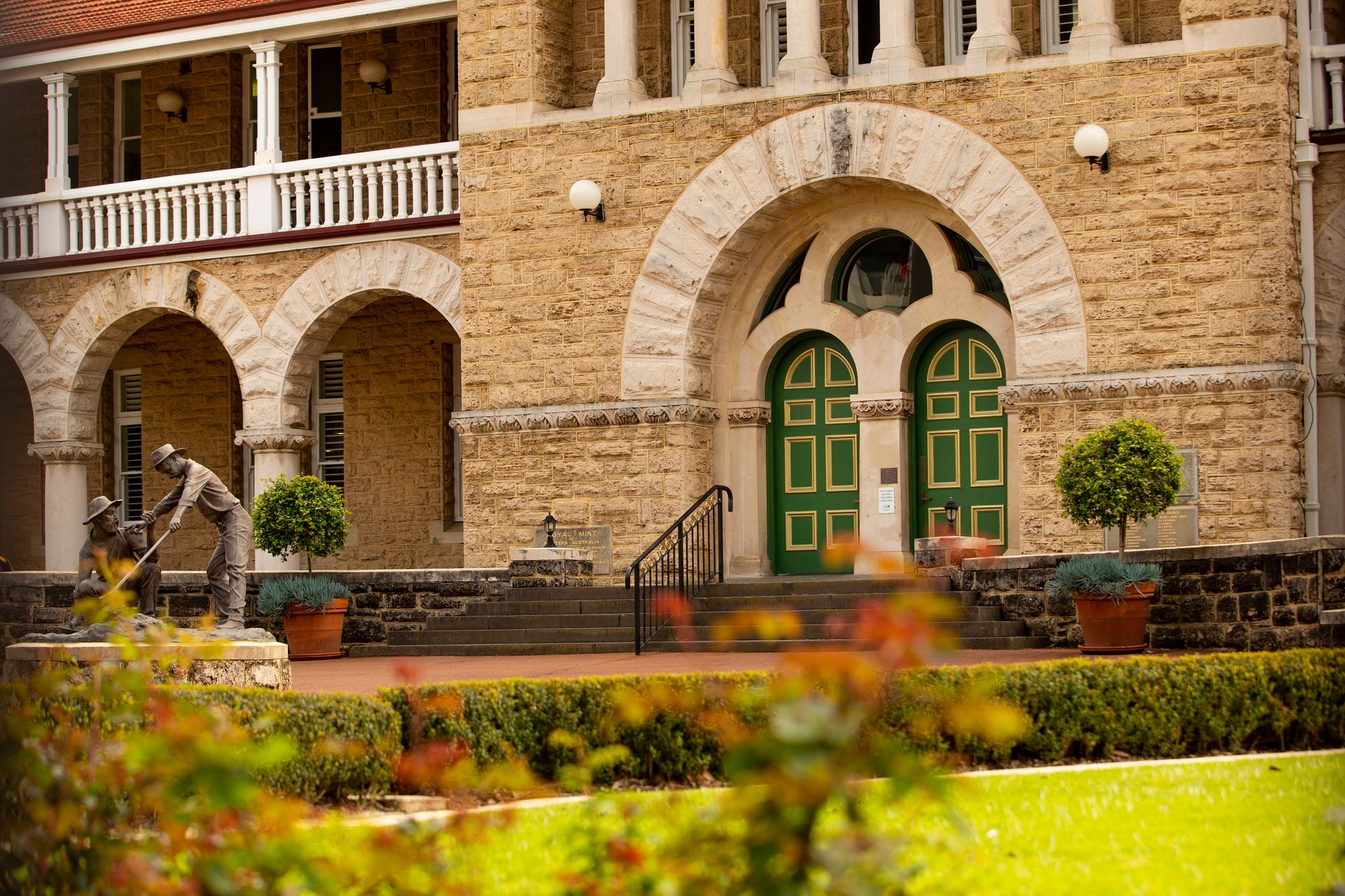 Garden featuring lawns, hedges and rose bushes in front of a large two storey building with limestone facade, orange tiled roof, arched colondade, two arched green with gold detail front doors.