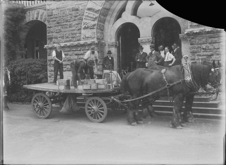 Black and white photo of arched double entrance doors to large building with a limestone facade; a horse drawn wagon is in front of the doors and men are unloading wooden boxes from it.