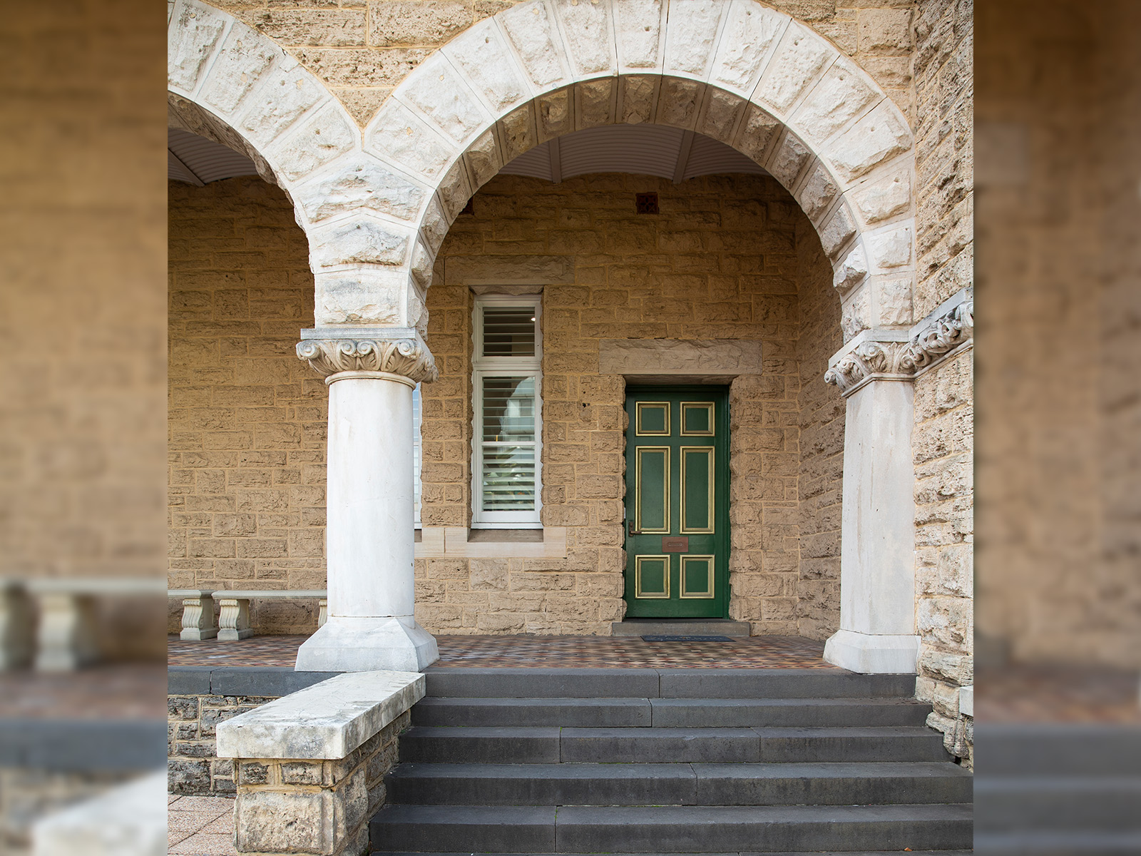 A rectangular green door recessed under an arched arcade of a building with a limestone facade. Flight of five steps leads up to the tiled arcade.