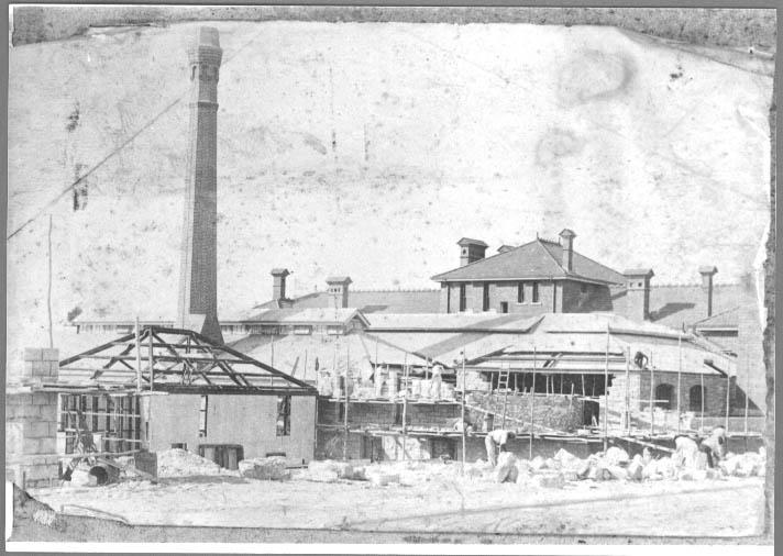 Black and white photo of brick factory buildings under construction. A tall brick chimney towers over other structures.