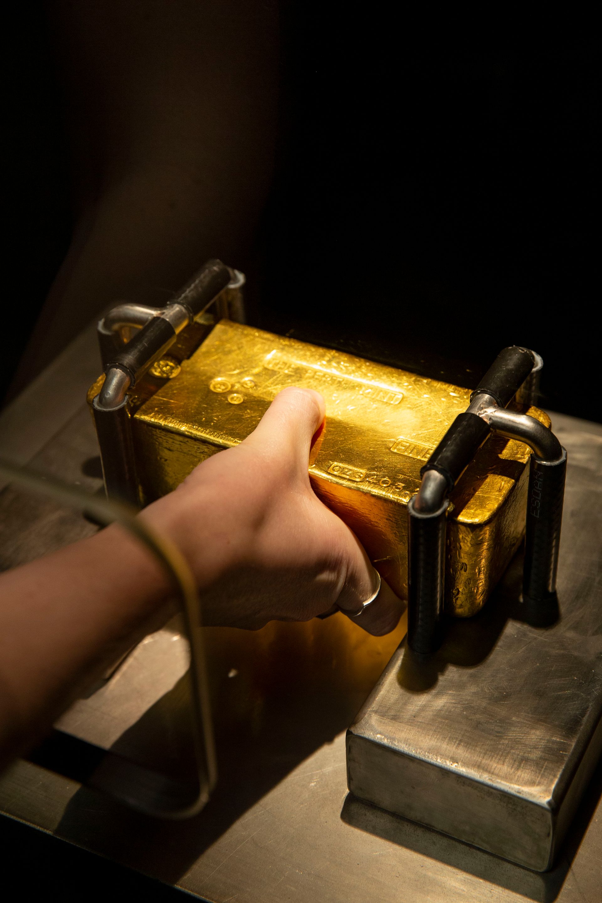 A hand extends through an opening in a display case to grasp the centre of a 12.4 kilogram cast gold bar to try and lift it as high as the metal restraints at both ends will allow