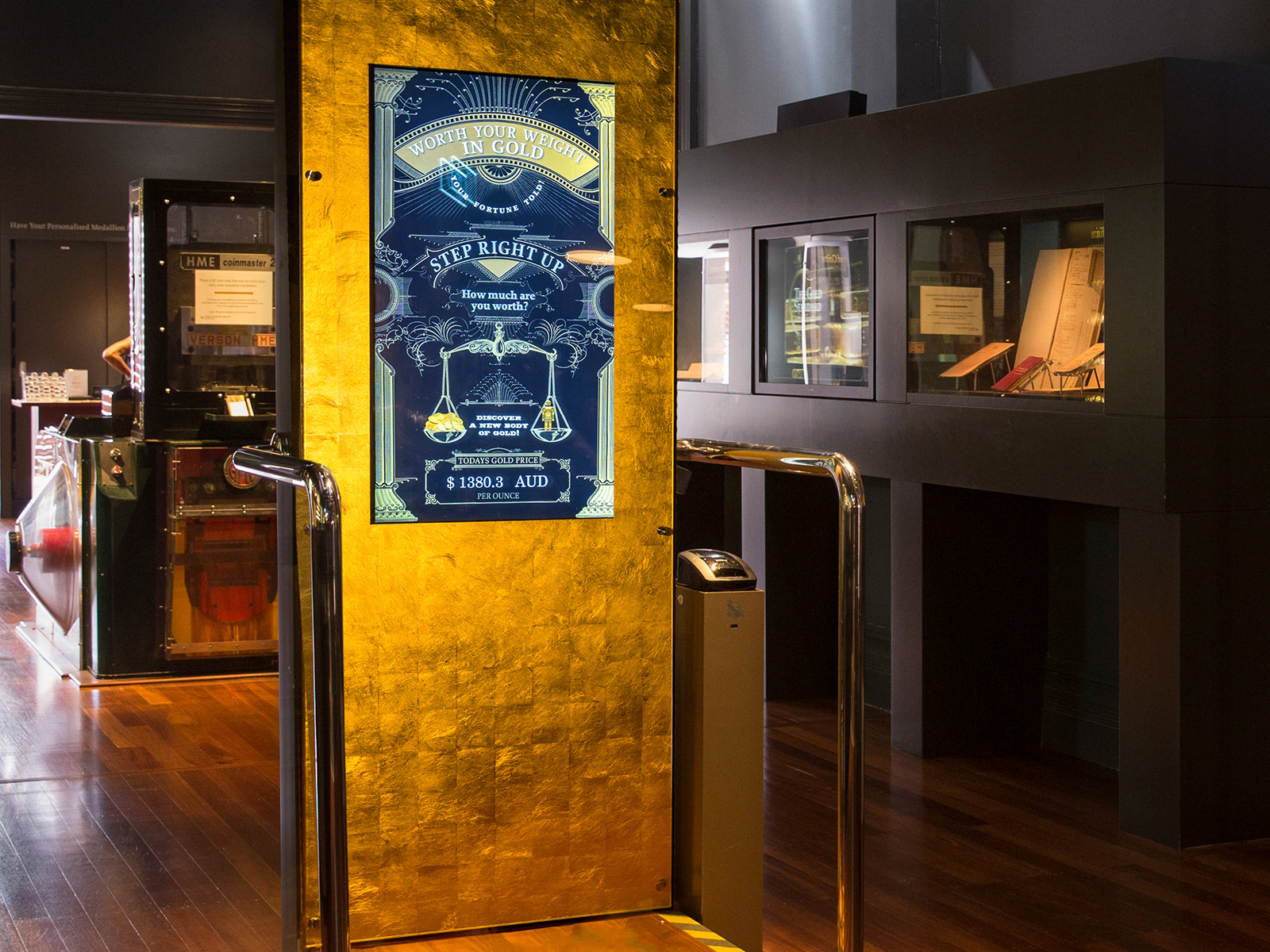 A set of scales with a tall gold panel with inset digital screen 'Worth Your Weight in Gold' stands in the centre of a museum gallery.