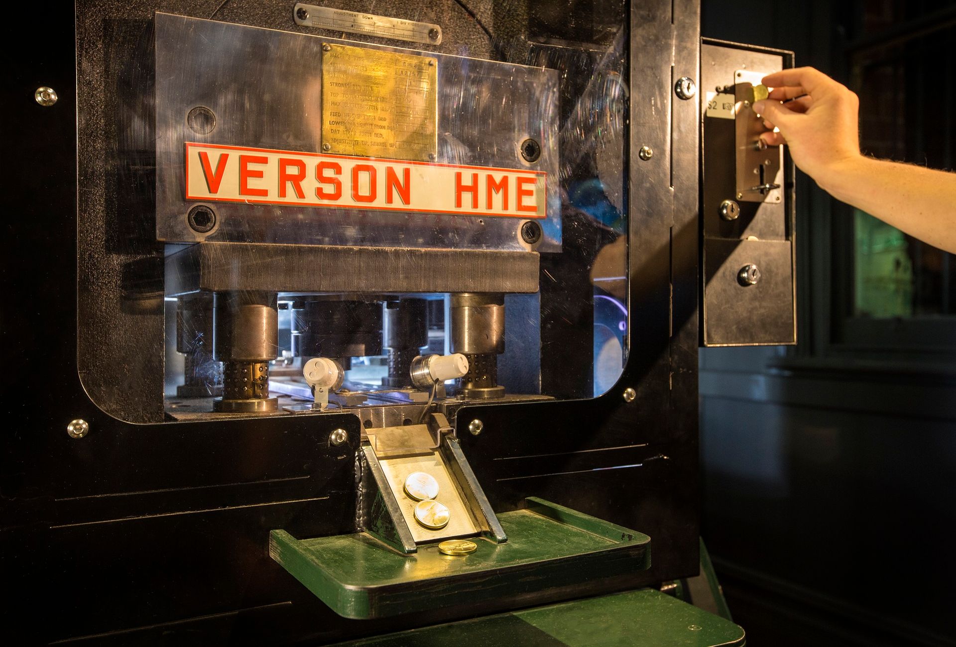 A hand is feeding a blank token into a slot attached to a coin press 'VERSON HME' and tokens struck with the design of The Perth Mint are sliding out down a chute.